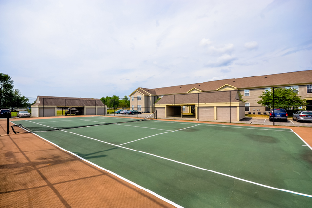 Empty outdoor tennis court with green and orange surface, surrounded by fencing, with apartment buildings and parked cars nearby