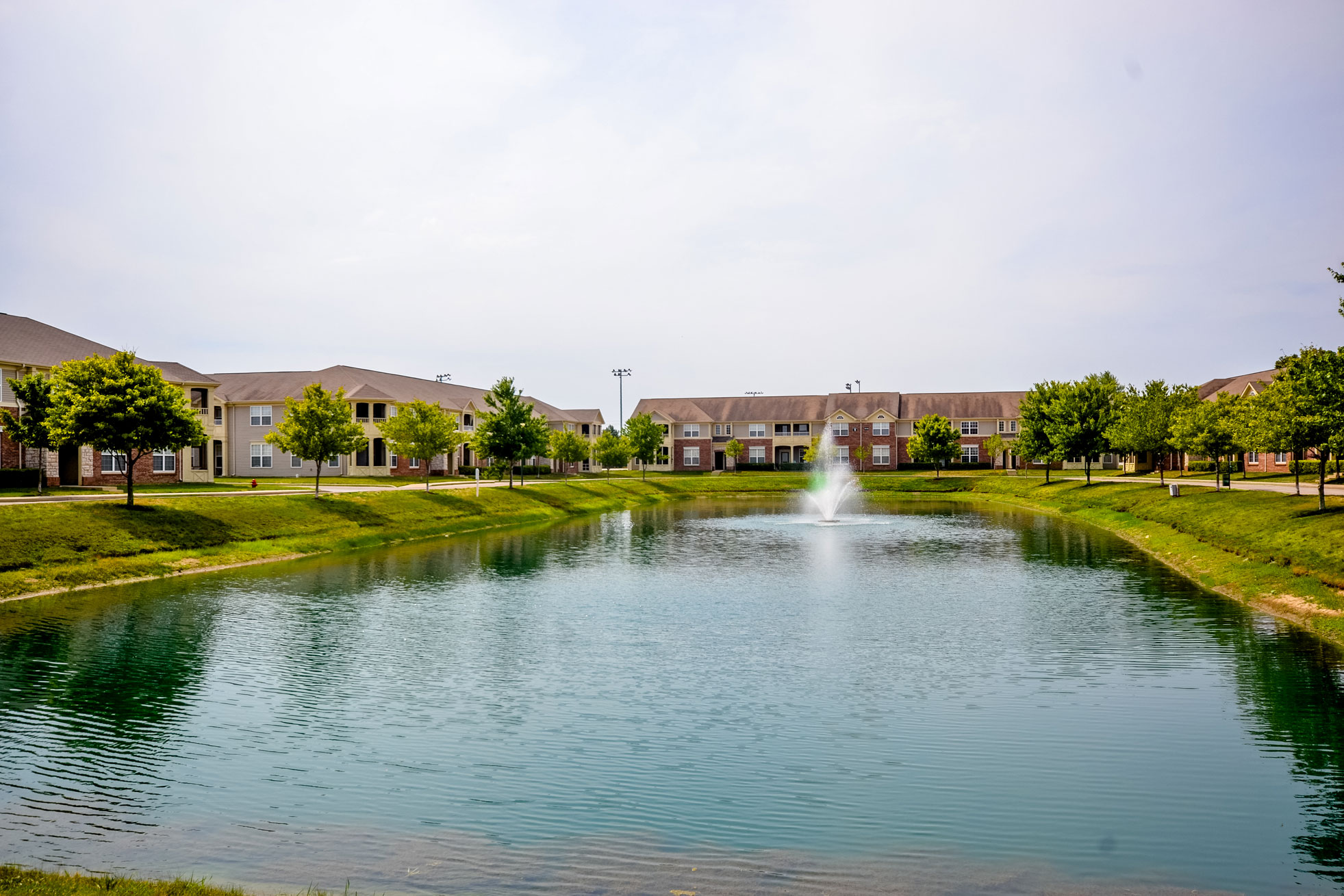 Pond with fountain surrounded by green lawn, trees, and multi-story residential buildings under a hazy sky